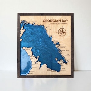 May include: Wooden wall art depicting a detailed map of Georgian Bay, Lake Huron, Ontario. The map is laser-cut into light-coloured wood, with blue epoxy resin representing the water. Includes a compass rose and text.