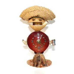 May include: A whimsical clock sculpture crafted from seashells, featuring a straw hat, a red clock face with Roman numerals, and a small can. The clock hands point to a specific time. The sculpture has a unique, handcrafted appearance.