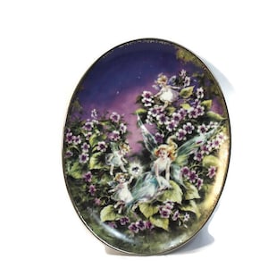 Peut inclure: Une assiette en porcelaine décorative avec un motif floral violet et vert représentant des fées avec des ailes et un bijou scintillant. L'assiette a un bord doré.