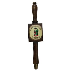May include: A wooden beer tap handle with a dark finish. The tap handle features a rectangular section with the text "CHARRINGTON TOBY" and an illustration of a man in green clothing. The handle is designed for dispensing beer.