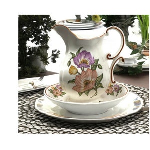 May include: A white ceramic pitcher with a floral design featuring purple, pink, and yellow flowers. The pitcher has a brown handle and spout, and sits atop a matching bowl and plate set. The set is ideal for serving beverages.