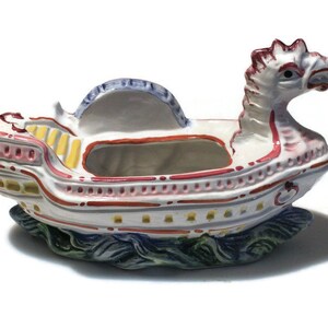 May include: A white ceramic planter shaped like a ship with a dragon head at the bow. The ship is decorated with red, yellow, and blue paint and has a small opening in the center for planting.