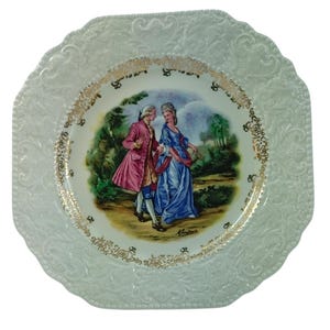 May include: A white square ceramic plate with a raised, embossed design around the edge. The center of the plate features a colorful image of a couple in a garden setting. The image is surrounded by a gold trim.