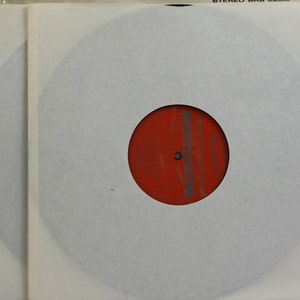 Peut inclure: Un disque vinyle dans une pochette en papier blanc. L'&eacute;tiquette du disque est rouge avec des lignes grises verticales. La pochette est &agrave; l'int&eacute;rieur d'une pochette ext&eacute;rieure en plastique transparent. Le haut de la pochette transparente porte l'inscription "STEREO SRS 02016".