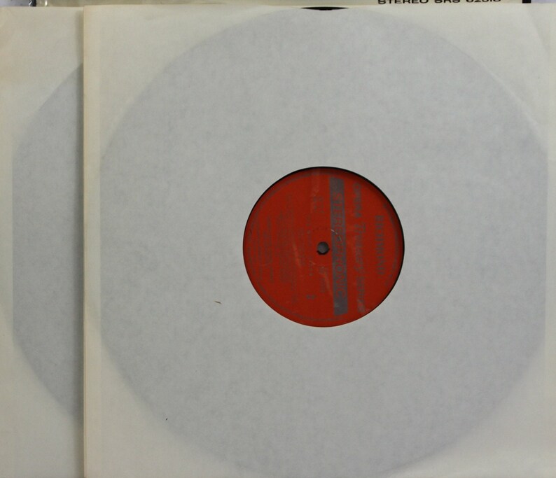 Peut inclure: Un disque vinyle dans une pochette en papier blanc. L'&eacute;tiquette du disque est rouge avec des lignes grises verticales. La pochette est &agrave; l'int&eacute;rieur d'une pochette ext&eacute;rieure en plastique transparent. Le haut de la pochette transparente porte l'inscription "STEREO SRS 02016".