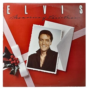 Puede incluir: Una portada de disco de vinilo rojo con el título "Elvis Memories of Christmas". La portada presenta una foto de Elvis Presley con una camisa negra sobre un fondo blanco.