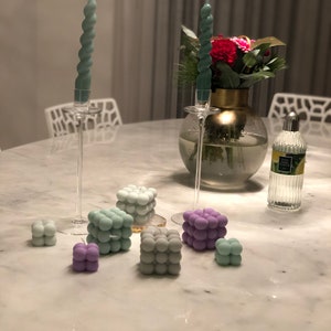 May include: A set of teal and purple bubble candles on a white marble table. The candles are arranged in a cluster, with two teal candles in the center and the purple candles surrounding them. The candles are all in the shape of a cube, with a textured surface that resembles bubbles.
