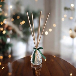 May include: A clear glass bottle shaped like a Christmas tree with a green ribbon and wooden diffuser sticks. The bottle is sitting on a wooden table.