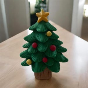 May include: A small, handcrafted Christmas tree figurine. The tree is a vibrant green with red and yellow ornaments. A yellow star tops the tree, and the base is a brown, textured trunk. The figurine is set on a light-colored wooden surface.