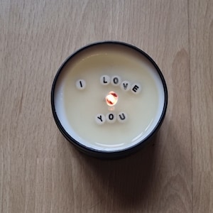 May include: A burning candle in a black tin. The cream-coloured wax has the words "I LOVE YOU" spelled out in small white letter beads. The flame is bright orange and is in the centre of the candle.