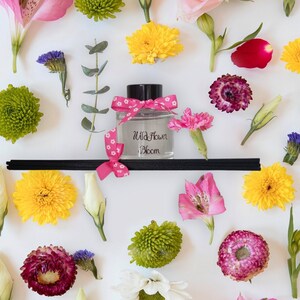 May include: A clear glass bottle with a black lid and pink ribbon, filled with a diffuser oil. The bottle is labelled "Wild Flower Bloom". The bottle is surrounded by various flowers, including yellow, pink, and green.
