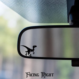 May include: A black vinyl decal of a velociraptor dinosaur silhouette applied to the surface of a vehicle rearview mirror. The dinosaur is depicted in a running pose, facing toward the right side of the mirror frame, with sharp claws visible.