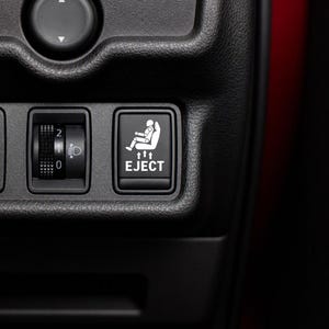 May include: A black rectangular car interior button featuring a white graphic of a person in a seat with three upward-pointing arrows and the text EJECT printed below. Next to it is a black headlight leveling dial with numbers 0, 1, and 2.