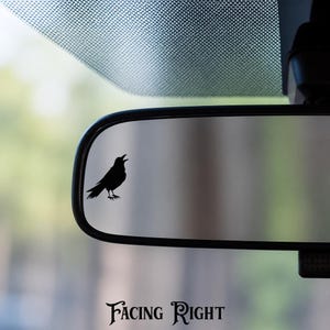 May include: A black silhouette decal of a crow with its beak open, positioned on the left side of a rectangular vehicle rearview mirror. The bird is depicted in profile, facing toward the right side of the mirror, with detailed feather markings.