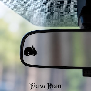 May include: A black silhouette decal of a rabbit in a seated position is applied to the surface of a vehicle rearview mirror. The rabbit is oriented toward the right side of the mirror. The text "FACING RIGHT" is printed below the mirror.