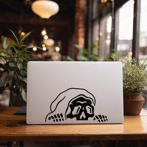 May include: A white laptop with a black graphic of a hooded skull. The skull is peering over the edge of the laptop, with its bony hands visible. The laptop is on a wooden table.