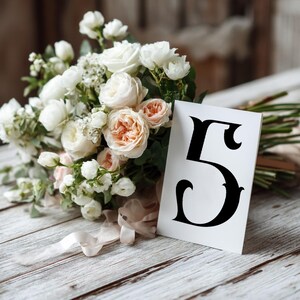 May include: A bouquet of white and peach roses and other flowers rests on a white wooden surface. A white card with the number 5 in black is propped up in front of the flowers. A light pink ribbon is tied around the stems.