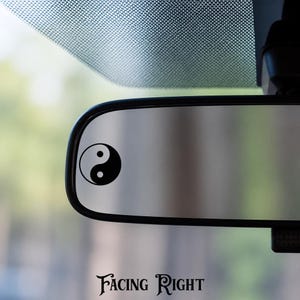 May include: A rectangular car rearview mirror featuring a black yin yang symbol decal applied to the left side of the reflective surface. The text "FACING RIGHT" is printed in a decorative black font below the mirror assembly.