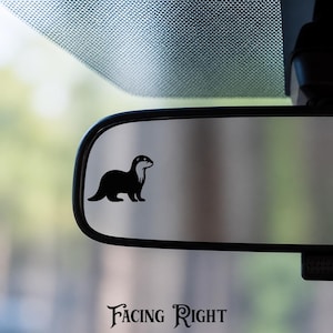 May include: A black silhouette decal of an otter facing right, applied to the surface of a vehicle rearview mirror. The otter is depicted in profile with a smooth, solid black finish, positioned on the left side of the reflective glass surface.