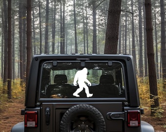 May include: A white Bigfoot silhouette decal is affixed to the rear window of a black SUV. The vehicle is parked on a dirt road in a forest with tall, slender trees and a misty background. The spare tire is mounted on the back.