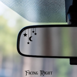 May include: A black vinyl decal applied to a car rearview mirror, featuring a celestial design of a crescent moon and four stars hanging from thin vertical lines. The graphic is positioned in the upper left corner of the mirror surface.