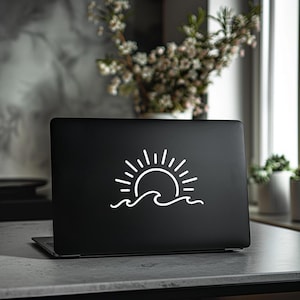 May include: Black laptop with a white graphic of a sun over ocean waves. The laptop is open on a gray surface. The background includes a vase of flowers and a window.