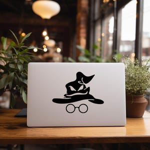 May include: A white laptop with a black sticker featuring a stylized image of the Sorting Hat and round glasses. The laptop is on a wooden table, with plants and a window in the background.