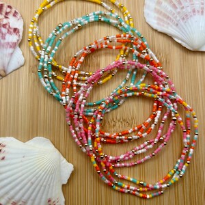 May include: A collection of colorful beaded bracelets in shades of yellow, turquoise, orange, pink, and white, accented with gold-colored beads. The bracelets are arranged on a wooden surface, with seashells in the background.