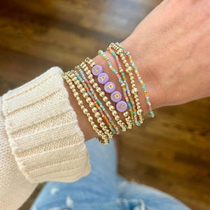May include: A collection of gold beaded bracelets and colorful seed bead bracelets. One bracelet features the word "HAPPY" spelled out in lavender letter beads. The bracelets are stacked together on a wrist.