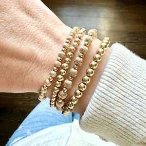 May include: Four gold beaded bracelets of varying designs are stacked on a wrist. The bracelets feature different bead styles, including smooth spheres and textured, ridged beads. The bracelets are a warm gold color and appear to be made of metal.