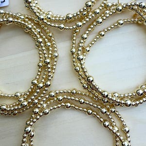 May include: A collection of circular bracelets and earrings crafted from polished gold-toned metallic beads of varying sizes. The jewelry pieces are arranged in a cluster, showcasing a reflective, smooth surface and a consistent spherical design.