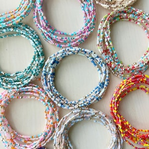 May include: A collection of colorful beaded bracelets in various shades, including turquoise, pink, blue, and yellow. Each bracelet is composed of small, colorful beads and interspersed with small, gold-colored beads. The bracelets are arranged in circular patterns.