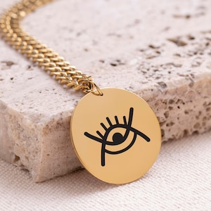 May include: Gold chain necklace with a round pendant featuring a black and white stylized eye with eyelashes.