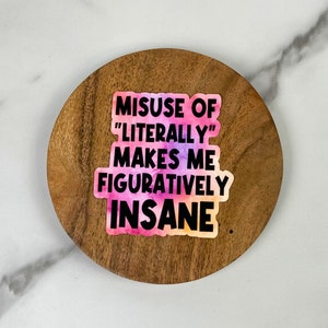 May include: A sticker with the text "MISUSE OF 'LITERALLY' MAKES ME FIGURATIVELY INSANE" in black font over a watercolor-style background of pink, purple, and orange. The sticker is on a round, wooden coaster.