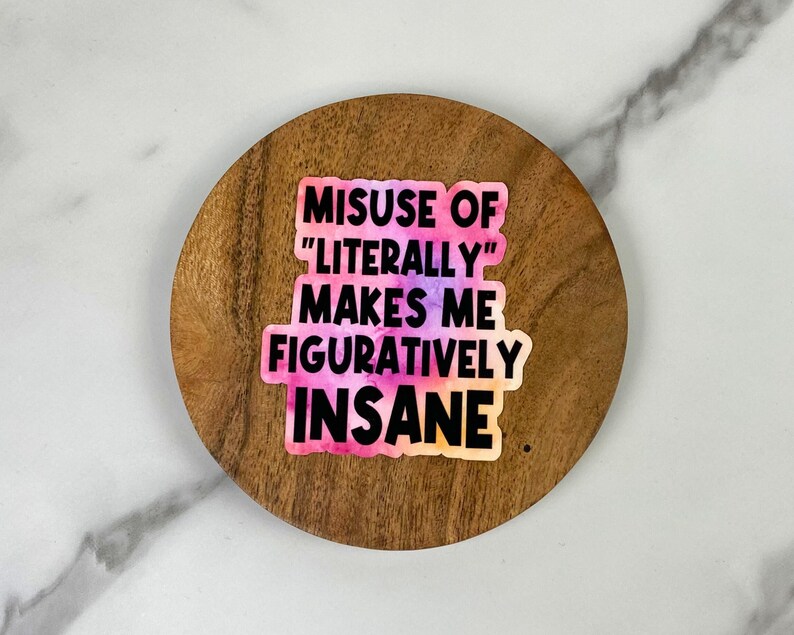 May include: A sticker with the text "MISUSE OF 'LITERALLY' MAKES ME FIGURATIVELY INSANE" in black font over a watercolor-style background of pink, purple, and orange. The sticker is on a round, wooden coaster.