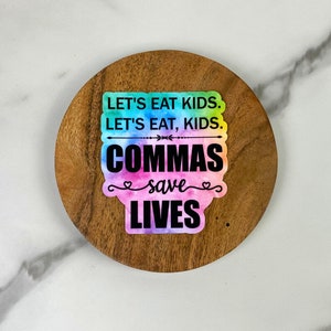 May include: A round, wooden coaster with a colorful sticker that reads "LET'S EAT KIDS. LET'S EAT, KIDS. COMMAS SAVE LIVES." The sticker has a tie-dye effect with blue, green, pink, and purple hues. The coaster has a natural wood grain.