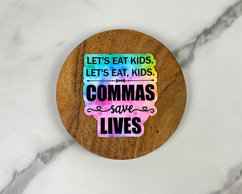 May include: A round, wooden coaster with a colorful sticker that reads "LET'S EAT KIDS. LET'S EAT, KIDS. COMMAS SAVE LIVES." The sticker has a tie-dye effect with blue, green, pink, and purple hues. The coaster has a natural wood grain.