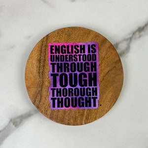 May include: A sticker with the text "ENGLISH IS UNDERSTOOD THROUGH TOUGH THOROUGH THOUGHT" in black and white, with a pink and purple gradient background. The sticker is on a round, wooden coaster.