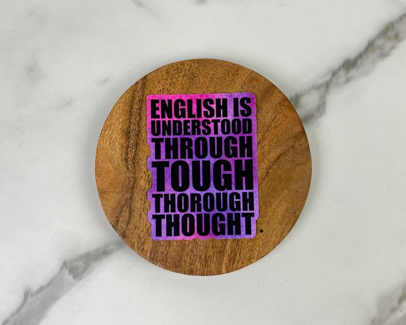 May include: A sticker with the text "ENGLISH IS UNDERSTOOD THROUGH TOUGH THOROUGH THOUGHT" in black and white, with a pink and purple gradient background. The sticker is on a round, wooden coaster.