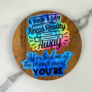 May include: A round wooden coaster with two colorful stickers. The top sticker reads "A BOOK A DAY Keeps Reality Away" with a rainbow watercolor effect. The bottom sticker says "You had me at the proper use of YOU'RE".