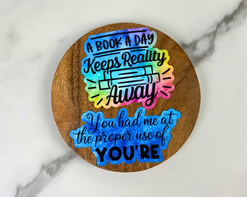 May include: A round wooden coaster with two colorful stickers. The top sticker reads "A BOOK A DAY Keeps Reality Away" with a rainbow watercolor effect. The bottom sticker says "You had me at the proper use of YOU'RE".