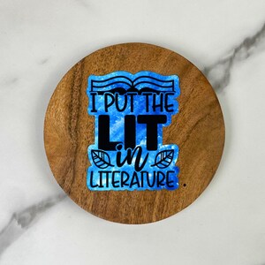 May include: A round wooden coaster with the text "I PUT THE LIT IN LITERATURE" in black and blue. The text is on a blue watercolor background with leaf accents. The coaster is made of a dark wood.