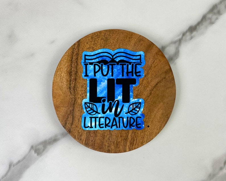 May include: A round wooden coaster with the text "I PUT THE LIT IN LITERATURE" in black and blue. The text is on a blue watercolor background with leaf accents. The coaster is made of a dark wood.