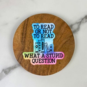 May include: A sticker with the text "TO READ OR NOT TO READ WHAT A STUPID QUESTION" and a watercolor design. The sticker features a stack of books in the center. The sticker is on a round wooden coaster.