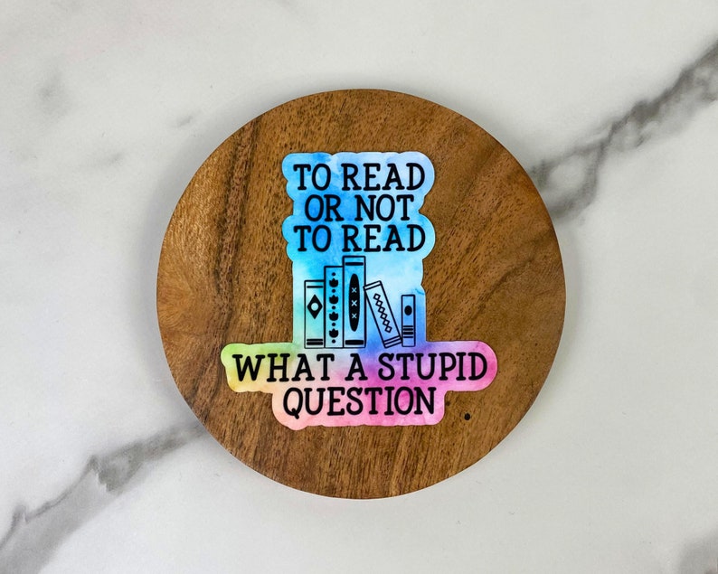 May include: A sticker with the text "TO READ OR NOT TO READ WHAT A STUPID QUESTION" and a watercolor design. The sticker features a stack of books in the center. The sticker is on a round wooden coaster.