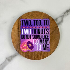 May include: A sticker with the text "TWO, TOO, TO TWO DONUTS DO NOT SOUND LIKE TOO MANY TO ME" in white and purple. Two cartoon donuts, one pink and one purple, are at the bottom. The sticker is on a wooden coaster.