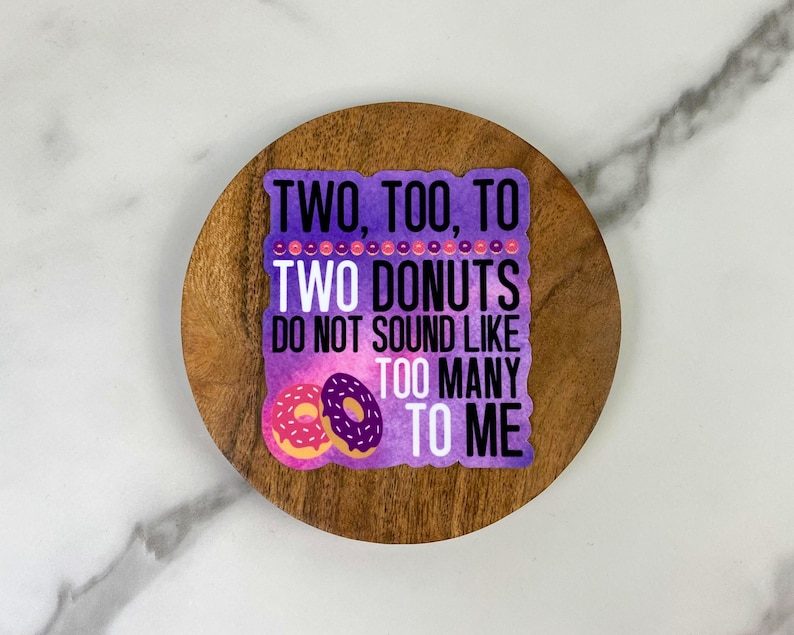 May include: A sticker with the text "TWO, TOO, TO TWO DONUTS DO NOT SOUND LIKE TOO MANY TO ME" in white and purple. Two cartoon donuts, one pink and one purple, are at the bottom. The sticker is on a wooden coaster.