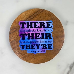 May include: A sticker with the words "THERE, THEIR, THEY'RE" in black, with a watercolor background of purple and blue. The sticker is on a wooden coaster, with the text "are people who didn't listen to teacher's grammar lessons, and driving me nuts!"