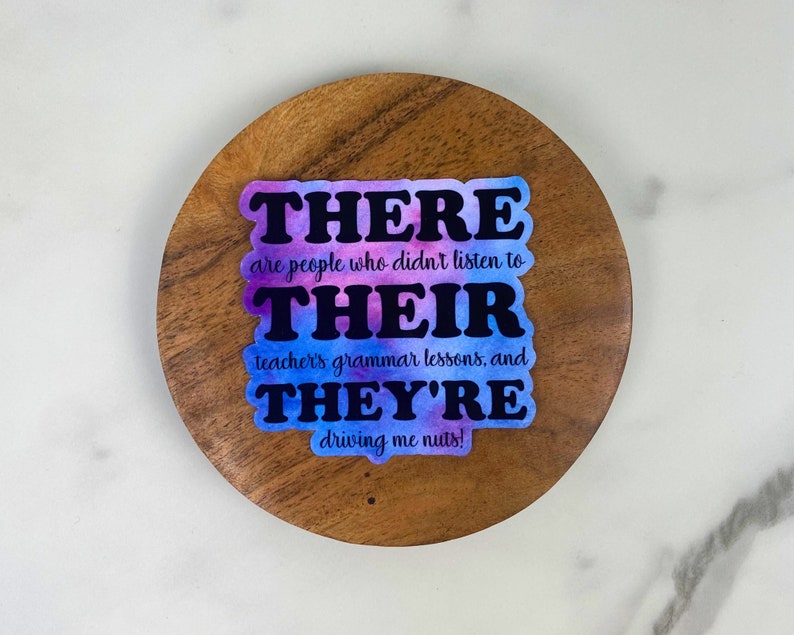 May include: A sticker with the words "THERE, THEIR, THEY'RE" in black, with a watercolor background of purple and blue. The sticker is on a wooden coaster, with the text "are people who didn't listen to teacher's grammar lessons, and driving me nuts!"