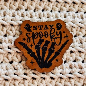 May include: A tan leather-like patch with a die-cut edge featuring a black graphic of a skeleton hand and two small ghosts. The text STAY SPOOKY is printed in black, accompanied by small star accents. The design is centered on a textured surface.